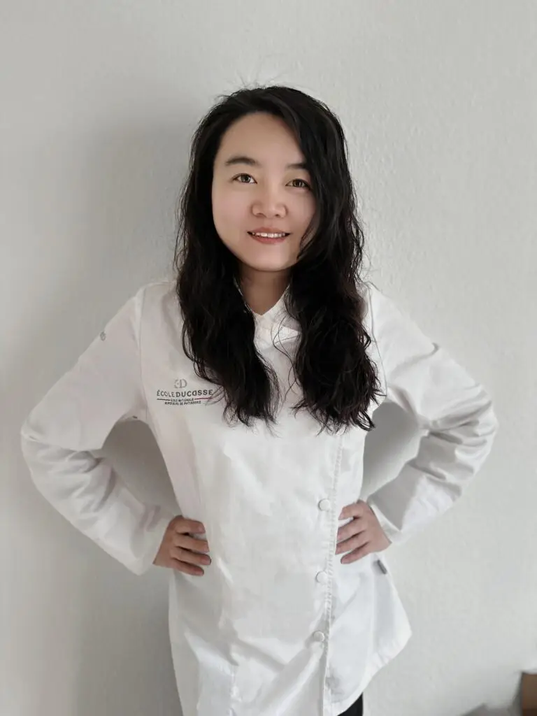 La cheffe pâtissière, debout en tenue de travail, qui vous regarde avec un sourire bienveillant
